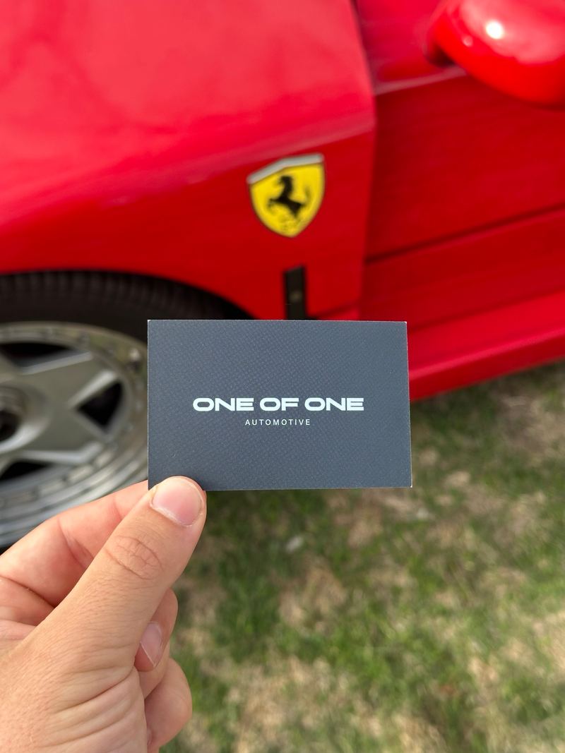 A One Of One Automotive business card held up against the red flank of a Ferrari.