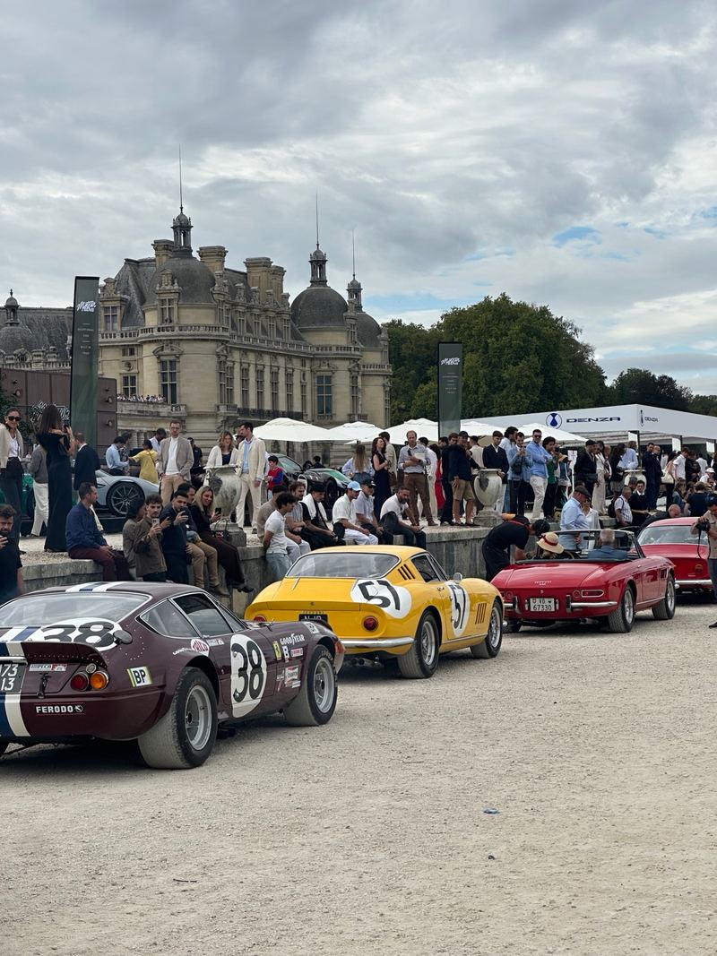 Classic Ferrari racing and road cars, including a yellow number 57 and a dark red number 38, lined up in front of the Château de Chantilly.