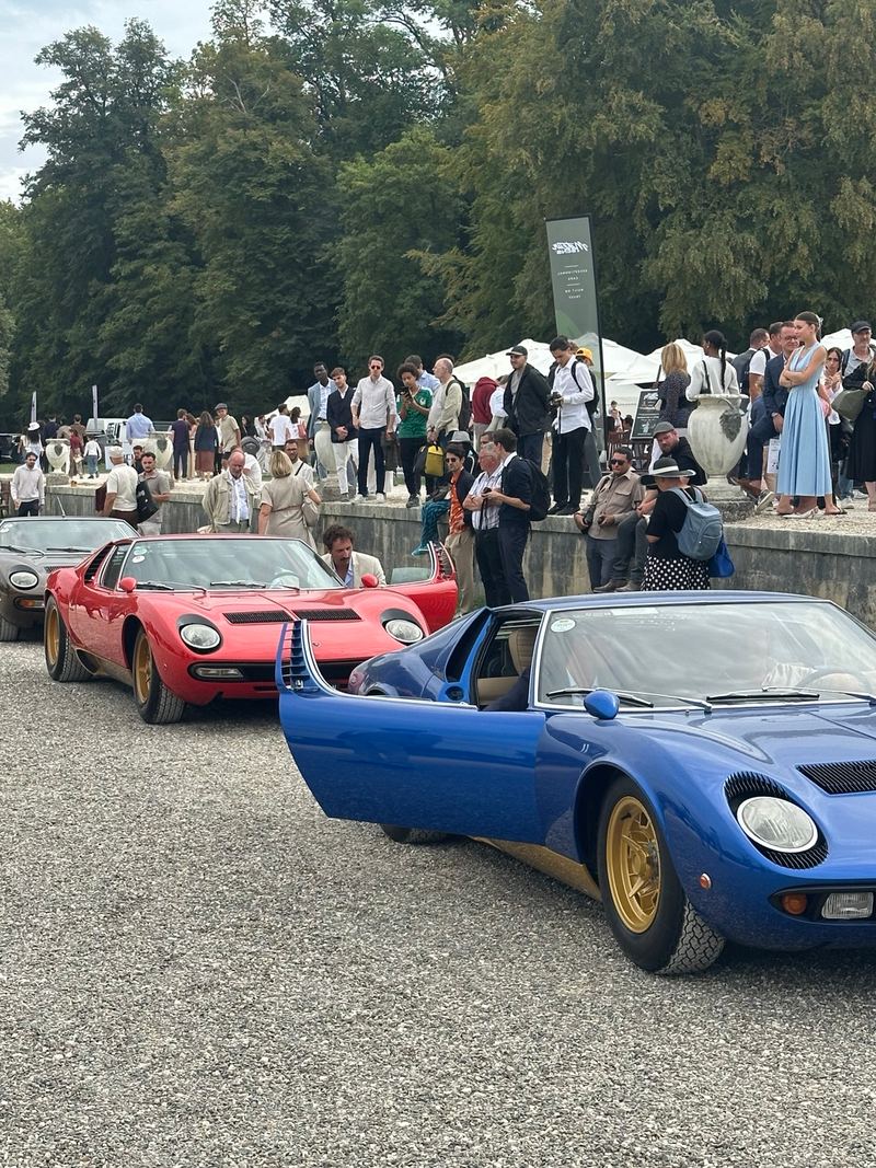 A blue and a red Lamborghini Miura on the gravel, the blue car’s door open, with spectators behind them.